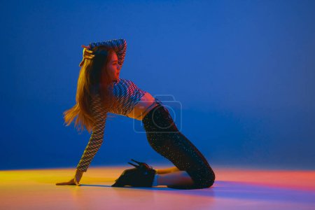 Foto de Portrait of young girl dancing high heel dance in stylish clothes over blue background in neon light. Concept of dance lifestyle, modern style, contemporary dance, youth culture, self-expression - Imagen libre de derechos