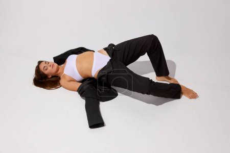 Téléchargez les photos : Portrait of young fit girl posing in white underwear and black classic suit over grey studio background. Concept of body and skin care, fitness, natural beauty, health, wellness. Copy space for ad - en image libre de droit