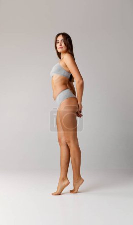 Téléchargez les photos : Fit. Portrait of young beautiful girl with slim body posing in cotton underwear over grey studio background. Concept of body and skin care, fitness, natural beauty, health, wellness. - en image libre de droit