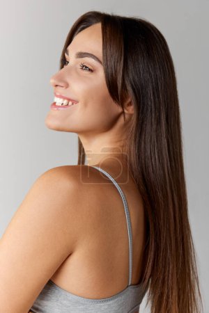 Téléchargez les photos : Portrait of young beautiful girl with straight brunette hair posing over grey studio background. Cosmetology care. Concept of body and skin care, fitness, natural beauty, health, wellness. - en image libre de droit