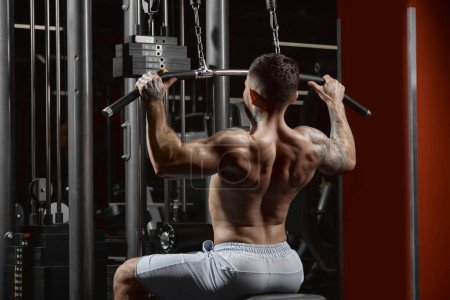 Téléchargez les photos : Young muscular man training shirtless in gym indoors. Doing lat pull down exercises. Side view. Relief, muscular back. Concept of health, sportive lifestyle, fitness, body care, diet, strength - en image libre de droit