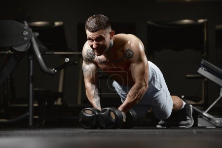 Téléchargez les photos : Endurance. Portrait of young muscular man training shirtless in gym indoors. Doing push ups with dumbbells. Concept of health, sportive lifestyle, fitness, body care, diet, strength - en image libre de droit