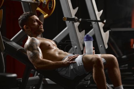 Photo for Portrait of young muscular man training shirtless in gym indoors. Resting after hard exercise session, drinking water. Concept of health, sportive lifestyle, fitness, body care, diet, strength - Royalty Free Image