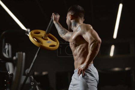Téléchargez les photos : Portrait of young muscular man training shirtless in gym indoors. Lifting barbell with one hand. Relief body, power. Concept of health, sportive lifestyle, fitness, body care, diet, strength - en image libre de droit