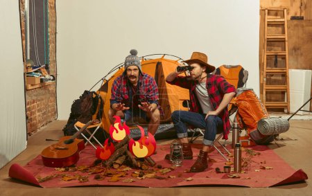 Foto de Looking around. Two men imagining, recreating camping activity indoors with necessary equipment. Concept of travelling, active lifestyle, friendship, leisure activity, relaxation - Imagen libre de derechos