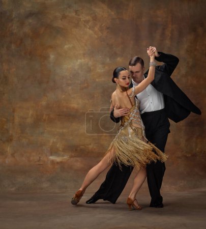 Téléchargez les photos : Man and woman, professional dancers in stylish stage costumes performing ballroom dance over dark vintage background. Coordinated movements. Concept of hobby, lifestyle, action, motion. - en image libre de droit