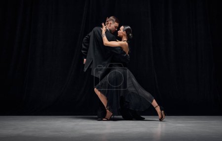 Photo for Passionate dance style. Man and woman, professional tango dancers performing in black stage costumes over black background. Concept of hobby, lifestyle, action, motion, art, dance aesthetics - Royalty Free Image