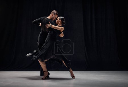 Téléchargez les photos : Man and woman, professional tango dancers performing in black stage costumes over black background. Expression of togetherness. Concept of hobby, lifestyle, action, motion, art, dance aesthetics - en image libre de droit