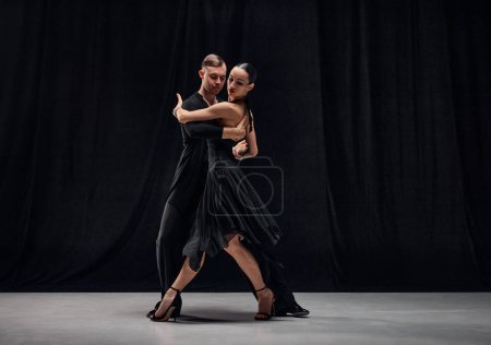 Photo for Style, passion, romance. Man and woman, professional tango dancers performing in black stage costumes over black background. Concept of hobby, lifestyle, action, motion, art, dance aesthetics - Royalty Free Image