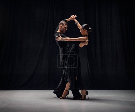 Photo for Deep attentive look. Man and woman, professional tango dancers performing in black stage costumes over black background. Concept of hobby, lifestyle, action, motion, art, dance aesthetics - Royalty Free Image