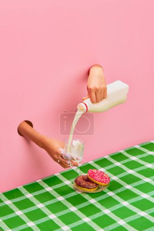 Téléchargez les photos : Food pop art photography. Female hands sticking out pink paper, pouring, spilling milk into cup. Donuts on green tablecloth. Concept of creativity, art. Complementary colors. Copy space for ad, text - en image libre de droit