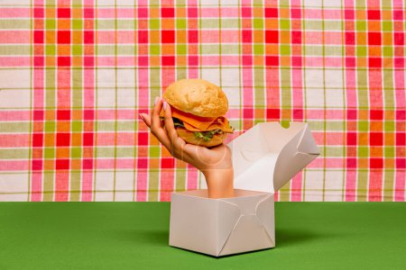 Téléchargez les photos : Food pop art photography. Female hand sticking out box and holding burger on green tablecloth. Junk food aesthetics. Concept of taste, creativity, art. Complementary colors. Copy space for ad, text - en image libre de droit