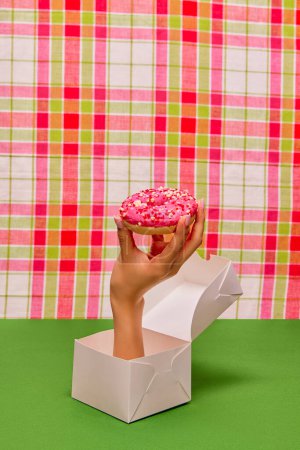 Téléchargez les photos : Food pop art photography. Female hand sticking out food box and holding donut with pink frosting on green tablecloth. Concept of taste, creativity, art. Complementary colors. Copy space for ad, text - en image libre de droit