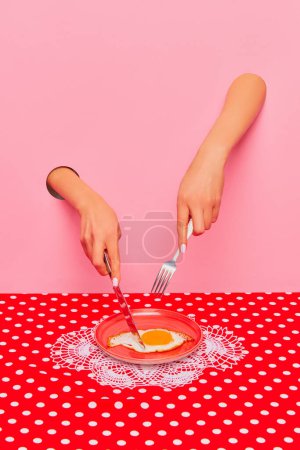 Téléchargez les photos : Food pop art photography. Female hand sticking out pink paper and cutting fried eggs on plate on retro tablecloth. Breakfast time. Taste, creativity, art. Complementary colors. Copy space for ad, text - en image libre de droit