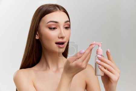 Téléchargez les photos : Young beautiful girl with perfect healthy skin applying gel, face cleanser over grey studio background. Concept of natural beauty, skin care, cosmetology, organic cosmetics, health, plastic surgery - en image libre de droit