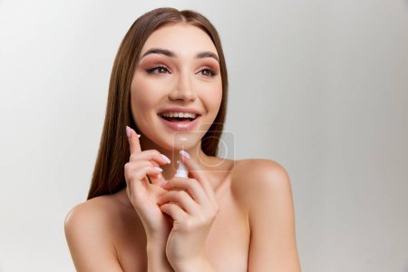 Téléchargez les photos : Smiling beautiful young girl with healthy, well-kept skin posing with face cream over grey studio background. Concept of natural beauty, skin care, cosmetology, cosmetics, health, plastic surgery - en image libre de droit