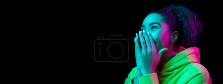 Téléchargez les photos : Positive shock. Young african girl covering mouth with hands in excitement and surprise over black background in neon light. Youth lifestyle. Concept of emotions, facial expression, sales, ad. Banner - en image libre de droit