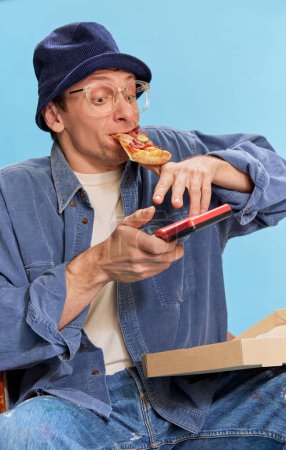 Téléchargez les photos : Leisure time. Cheerful man in jeans clothes sitting with pizza and playing retro game console over blue studio background. Concept of emotions, facial expression, lifestyle, retro fashion. Ad - en image libre de droit