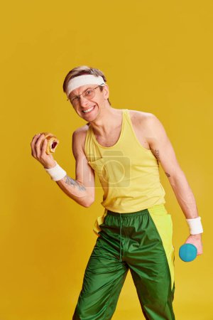 Téléchargez les photos : Portrait of young cheerful man in sports uniform with croissant and dumbbell over bright yellow studio background. Concept of emotions, facial expression, sportive lifestyle, retro fashion. Ad - en image libre de droit