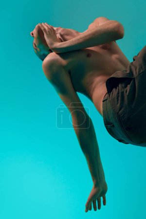Foto de Bottom view. Contemporary dance style. Young flexible man dancing over blue, cyan studio background. Art of movements, feelings. Concept of art, body aesthetics, motion, action, inspiration. - Imagen libre de derechos