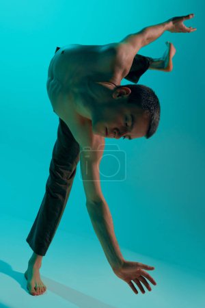 Téléchargez les photos : Contemporary dance style. Young flexible shirtless man dancing experimental dance over blue, cyan studio background. Artistic. Concept of art, body aesthetics, motion, action, inspiration. - en image libre de droit