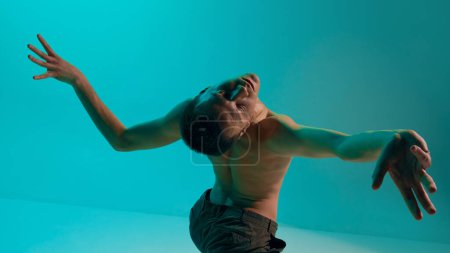 Photo for Bottom view. Contemporary dance style. Young expressive, flexible, shirtless man dancing over blue, cyan background. Experimental dance. Concept of art, body aesthetics, motion, action, inspiration - Royalty Free Image