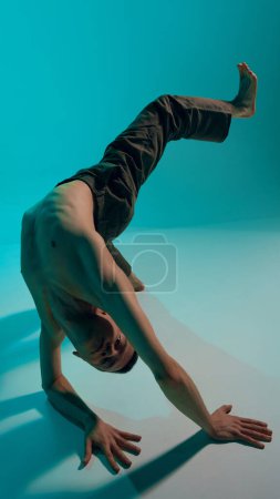 Foto de Contemporary dance style. Young flexible shirtless man dancing experimental dance over blue, cyan studio background. Active movements. Concept of art, body aesthetics, motion, action, inspiration. - Imagen libre de derechos