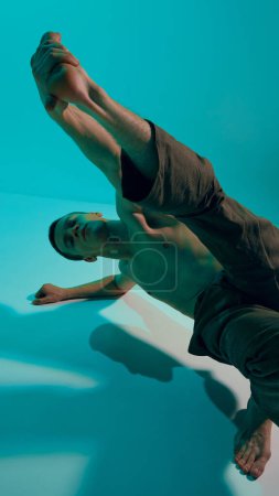 Photo for Bottom view. Contemporary dance style. Young expressive, flexible man dancing over blue, cyan studio background. Experimental dance. Concept of art, body aesthetics, motion, action, inspiration. - Royalty Free Image