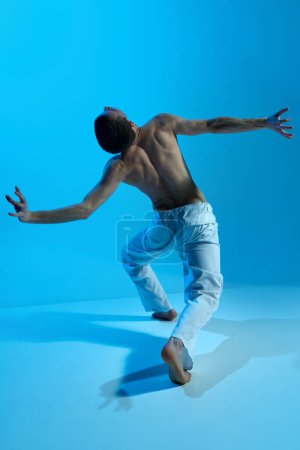 Téléchargez les photos : Contemporary dance style. Young artistic man dancing contemp, experimental dance over blue studio background. Beauty of movements. Concept of art, body aesthetics, motion, action, inspiration. - en image libre de droit