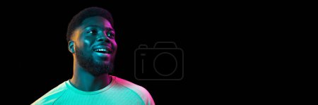 Téléchargez les photos : Positive vibes. Handsome young african man smiling, posing with excitement over black background in neon light. Banner, flyer. Concept of emotions, facial expression, sales, ad, fashion - en image libre de droit