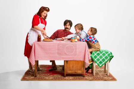 Téléchargez les photos : Noisy lovely family morning. Beautiful woman serving pie to her husband and sons sitting at kitchen table. Domestic lifestyle. Concept of retro style, family, 50s, 60s, fashion, childhood - en image libre de droit