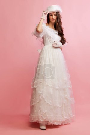 Téléchargez les photos : Portrait of beautiful lady in white vintage dress and hat on pink background. Concept of 19th century, fashion, comparison of eras, history, retro style. Female model in image of historical character - en image libre de droit