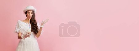 Foto de Celebration. Portrait of beautiful lady in white vintage dress posing with birthday cake over pink background. Concept of 19th century, fashion, comparison of eras, history, retro style - Imagen libre de derechos