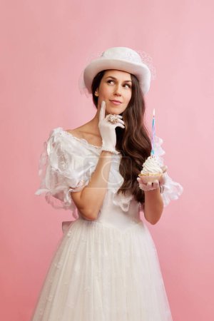 Foto de Making wishes. Portrait of beautiful lady in white vintage dress posing with birthday cupcake over pink background. Concept of 19th century, fashion, comparison of eras, history, retro style - Imagen libre de derechos