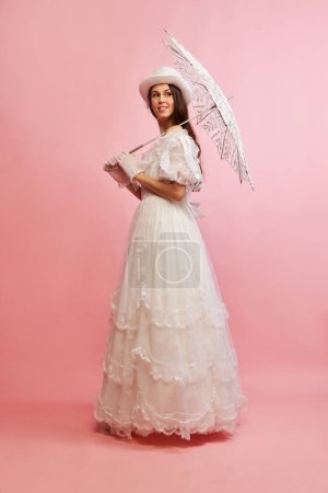 Tender femininity. Portrait of beautiful lady in white vintage dress posing with umbrella over pink background. Concept of 19th century, fashion, comparison of eras, history, retro style