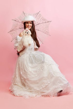 Photo for Lovely walk. Portrait of beautiful lady, woman in white vintage dress posing with cute dog over pink background. Concept of 19th century, fashion, comparison of eras, history, retro style - Royalty Free Image