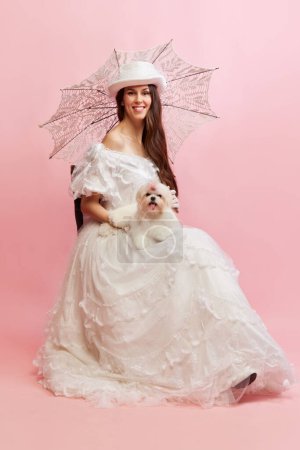 Foto de Woman and pet. Smiling. Portrait of beautiful lady in white vintage dress posing with dog. Concept of 19th century, fashion, comparison of eras, history. Female model in image of historical character - Imagen libre de derechos