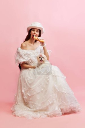 Foto de Eating hotdog. Portrait of beautiful lady in white vintage dress posing with dog over pink background. Fast food. Concept of 19th century, fashion, comparison of eras, history, retro style - Imagen libre de derechos