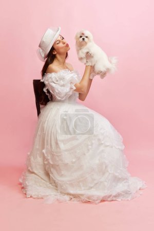 Foto de Portrait of beautiful lady, woman in white vintage dress posing with lovely cute dog over pink background. Concept of 19th century, fashion, comparison of eras, history, retro style - Imagen libre de derechos