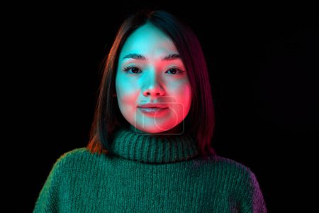 Photo for Looking at camera. Young beautiful girl with short brunette hair in cozy sweater over dark background in neon light. Concept of emotions, facial expression, youth, inspiration, sales, ad - Royalty Free Image
