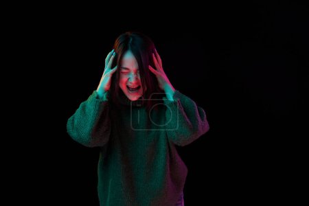 Photo for Anger, irritation, stress. Young girl in cozy sweater emotionally posing screaming over dark background in neon light. Concept of emotions, facial expression, youth, inspiration, sales, ad - Royalty Free Image