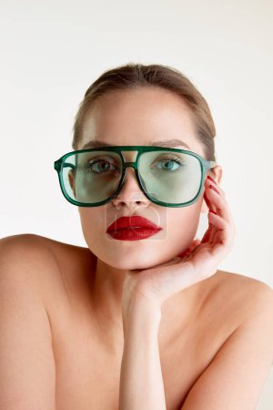 Téléchargez les photos : Femininity and sensuality. Young beautiful girl with red lips posing in green sunglases over grey studio background. Concept of natural beauty, skin care, cosmetology, cosmetics, health, fashion - en image libre de droit