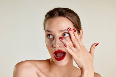 Téléchargez les photos : Portrait of young beautiful brunette girl with red lipstick and red nails posing over grey studio background. Well-kept skin. Concept of natural beauty, skin care, cosmetology, cosmetics, health - en image libre de droit