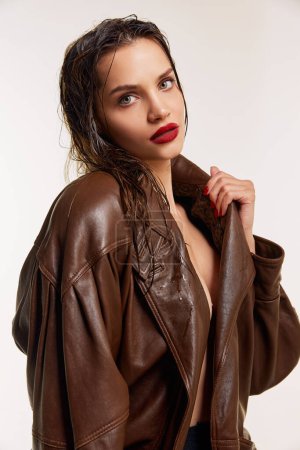 Téléchargez les photos : Attractive young brunette girl with wet hair and red lips in stylish leather jacket posing over grey studio background. Concept of natural beauty, skin care, cosmetology, cosmetics, modern fashion - en image libre de droit