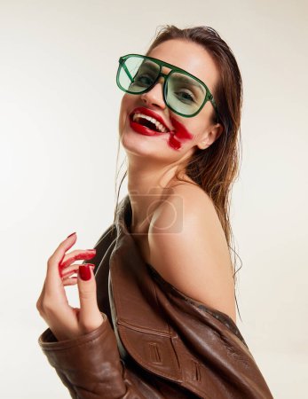 Téléchargez les photos : Red lipstick rubbed over. Beautiful young brunette girl with wet hair in stylish leather jacket smiling on grey studio background. Concept of natural beauty, skin care, cosmetology, cosmetics, fashion - en image libre de droit
