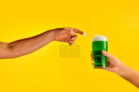 Téléchargez les photos : Male hand reaching glass with green foamy frothy beer over yellow background. Concept of st patricks day celebration, brewery, traditions, alcohol drinks, taste, Irish holiday - en image libre de droit