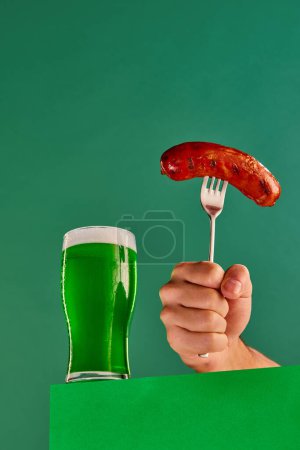 Foto de Glass with foam green beer and make hand with sausage on fork on green background. Traditional food and drink combination. Concept of st patricks day celebration, alcohol drinks, taste, Irish holiday - Imagen libre de derechos