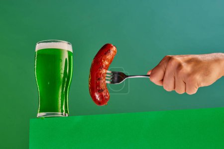 Photo for Glass with foam green beer and make hand with sausage on fork on green background. Traditional food and drink combination. Concept of st patricks day celebration, alcohol drinks, taste, Irish holiday - Royalty Free Image
