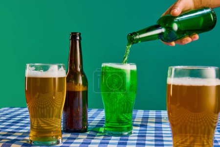 Foto de Bottle and glasses with lager and green foamy beer on checkered tablecloth over green background. Concept of st patricks day celebration, brewery, traditions, alcohol drinks, taste, Irish holiday - Imagen libre de derechos