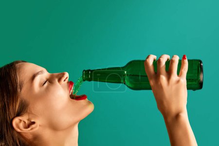 Téléchargez les photos : Woman pouring green beer into her mouth over green studio background. Concept of st patricks day celebration, brewery, traditions, alcohol drinks, taste, Irish holiday - en image libre de droit